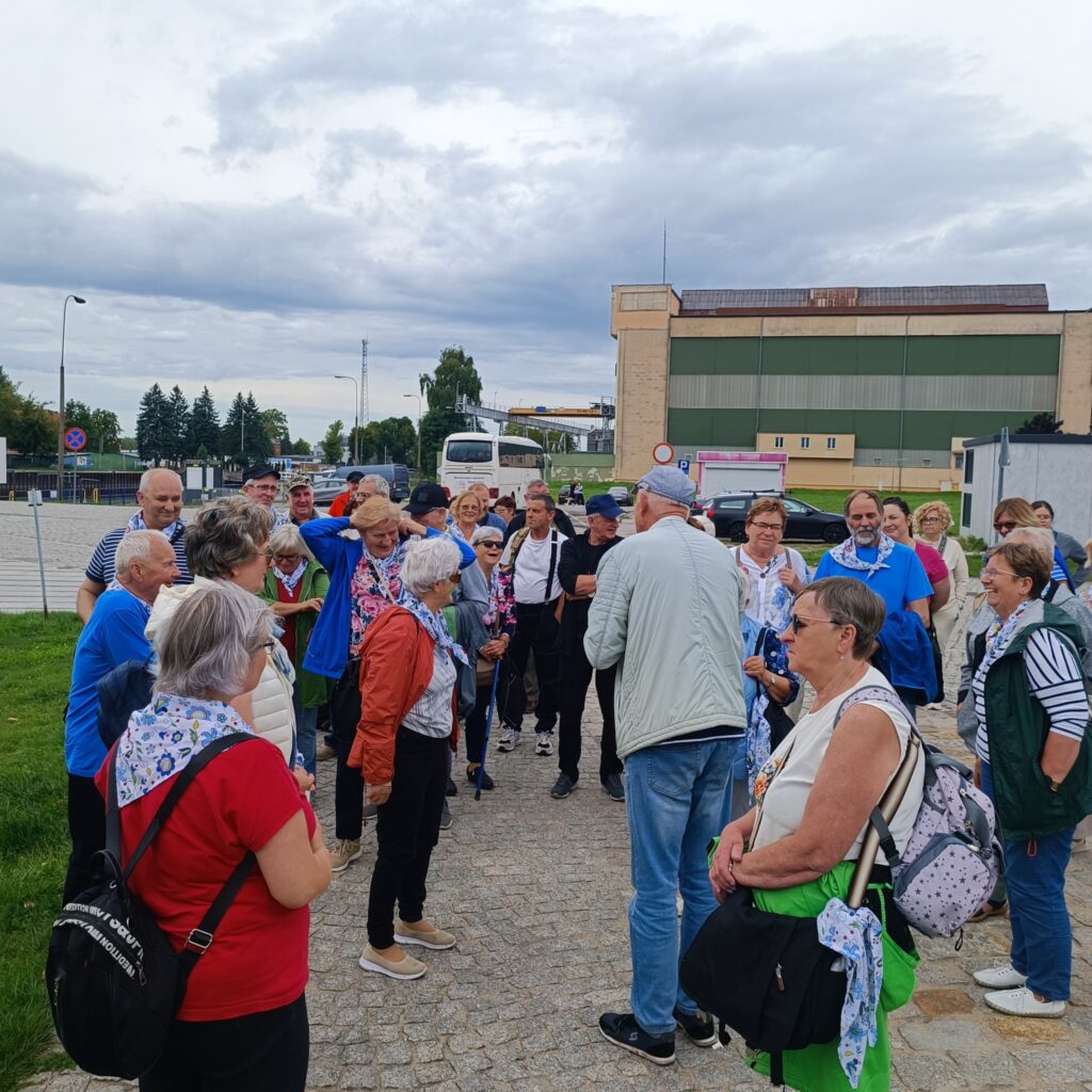 Grupa uczestników wyjazdu rehabilitacyjnego w trakcie spotkania przed rozpoczęciem zwiedzania w Elblągu. Uczestnicy w różnym wieku, w tym seniorzy, są ubrani w kolorowe chusty i aktywnie przygotowują się do wspólnej wyprawy. W tle widoczny jest autokar, który przewoził uczestników na wycieczkę. Niezwykle miła i zaangażowana atmosfera, gotowa do podjęcia kolejnych wyzwań.