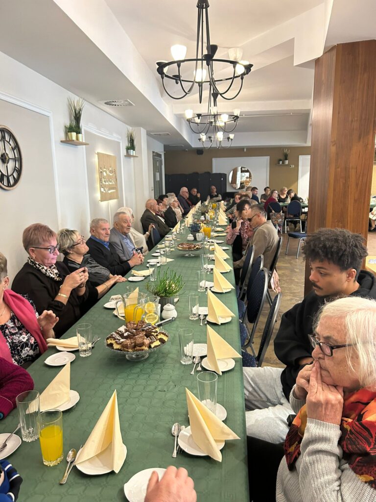 Zdjęcie przedstawia długi stół ustawiony w restauracji „Kociewianka” podczas uroczystości z okazji Dnia Białej Laski. Uczestnicy spotkania siedzą po obu stronach stołu, a na stole znajdują się filiżanki, szklanki, napoje oraz talerze z ciastem. Goście, w tym osoby starsze, są skupieni na rozmowie i wydarzeniach, niektórzy z nich robią zdjęcia lub słuchają uważnie. W tle widać eleganckie wnętrze lokalu, oświetlone klasycznymi żyrandolami. Cała scena ma spokojną, wspólnotową atmosferę.