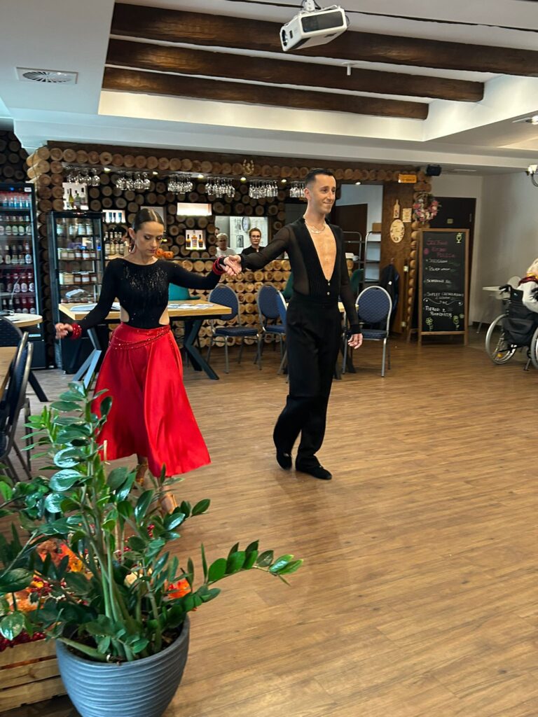 Na zdjęciu widoczna jest para taneczna, która występuje na parkiecie restauracji „Kociewianka” podczas uroczystości z okazji Dnia Białej Laski. Kobieta w czerwonej spódnicy i czarnej bluzce z cekinami tańczy w parze z mężczyzną, który ma na sobie czarny strój taneczny. Oboje trzymają się za ręce, wykonując elegancki taniec. W tle widać gości siedzących przy stolikach oraz dekoracyjną ścianę z okrągłych desek. Cała scena ma dynamiczną i radosną atmosferę, a tańcząca para przyciąga uwagę zgromadzonych.