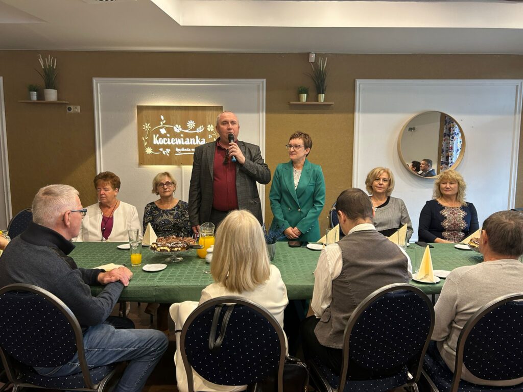 Na zdjęciu widoczny jest mężczyzna przemawiający do zgromadzonych osób przy stole w restauracji „Kociewianka” z okazji Dnia Białej Laski. Mężczyzna trzyma mikrofon i zwraca się do gości, a obok niego stoi kobieta w zielonym garniturze, słuchając uważnie. W tle widać grupę uczestników spotkania, którzy siedzą przy stole, w tym kilka kobiet, które patrzą na mówiącego. Na stole znajdują się napoje i kawałki ciasta, a cała scena odbywa się w jasnej, przytulnej sali z dekoracyjnymi elementami. W tle na ścianie widoczny jest napis „Kociewianka” oraz okrągłe lustro odbijające fragment wnętrza. Atmosfera spotkania jest serdeczna i uroczysta.