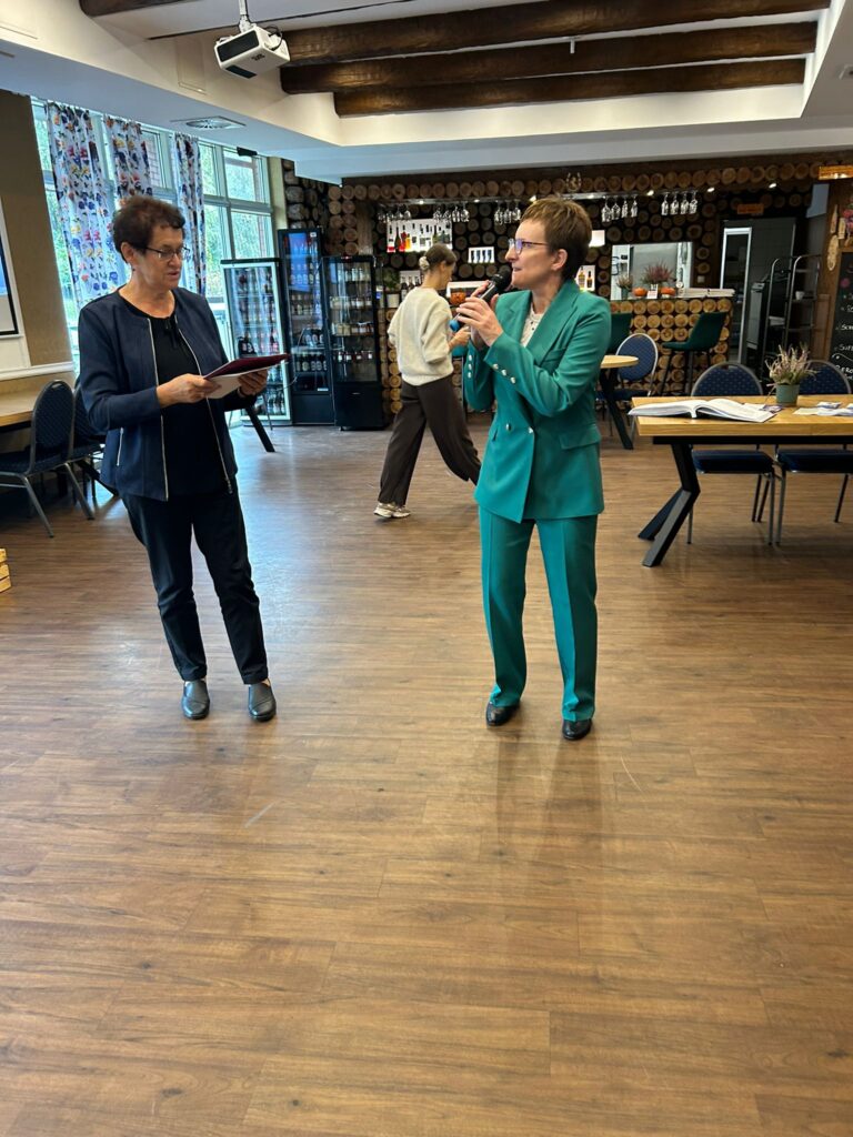 Na zdjęciu widać dwie kobiety stojące w centrum sali restauracji „Kociewianka” podczas uroczystości z okazji Dnia Białej Laski. Jedna z nich trzyma w ręku dokumenty i patrzy na drugą, która trzyma mikrofon i przemawia. Kobieta z mikrofonem jest ubrana w zielony garnitur, a druga w ciemną marynarkę. W tle widać drewnianą ścianę z okrągłymi deskami, regały z napojami oraz stolik z materiałami informacyjnymi. W oddali widoczna jest osoba, która przechodzi przez salę. Cała scena ma profesjonalną, lecz przyjazną atmosferę.