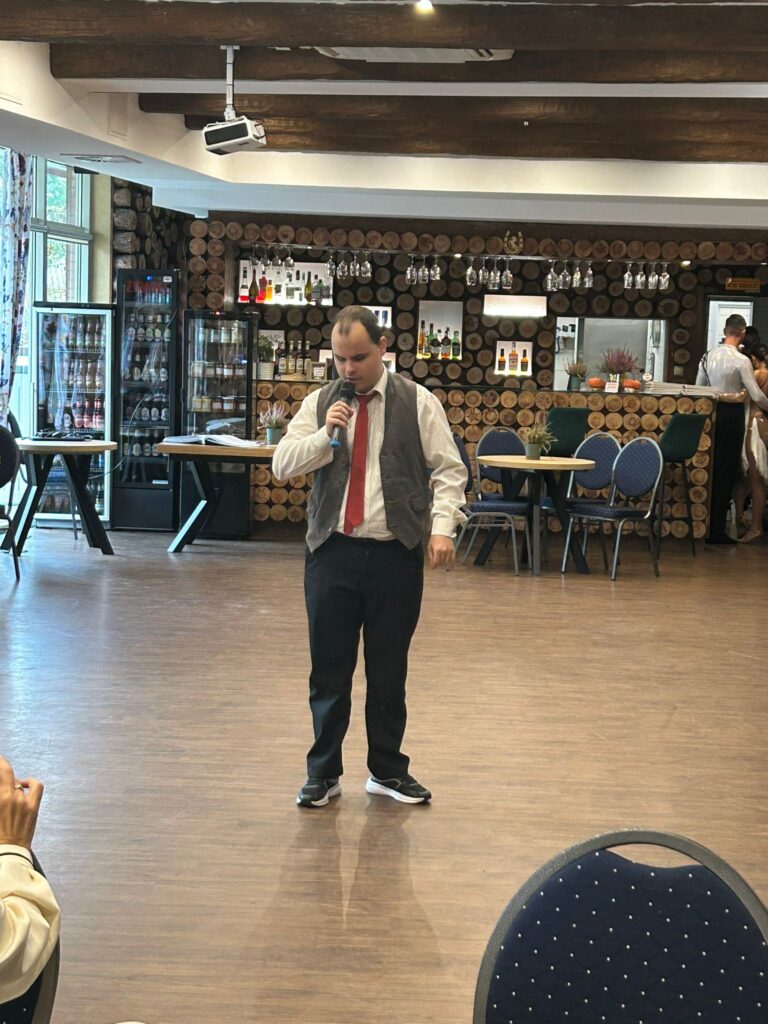 Zdjęcie przedstawia mężczyznę stojącego na środku sali restauracji „Kociewianka” podczas uroczystości z okazji Dnia Białej Laski. Mężczyzna trzyma mikrofon i śpiewa lub przemawia, ubrany w białą koszulę, szary kamizelkę, czarne spodnie i sportowe buty. W tle widać stylowe wnętrze restauracji z drewnianą ścianą z okrągłych desek, na której wiszą szklane kieliszki i butelki. Sala jest przestronna, a atmosfera jest radosna i pełna zaangażowania gości.