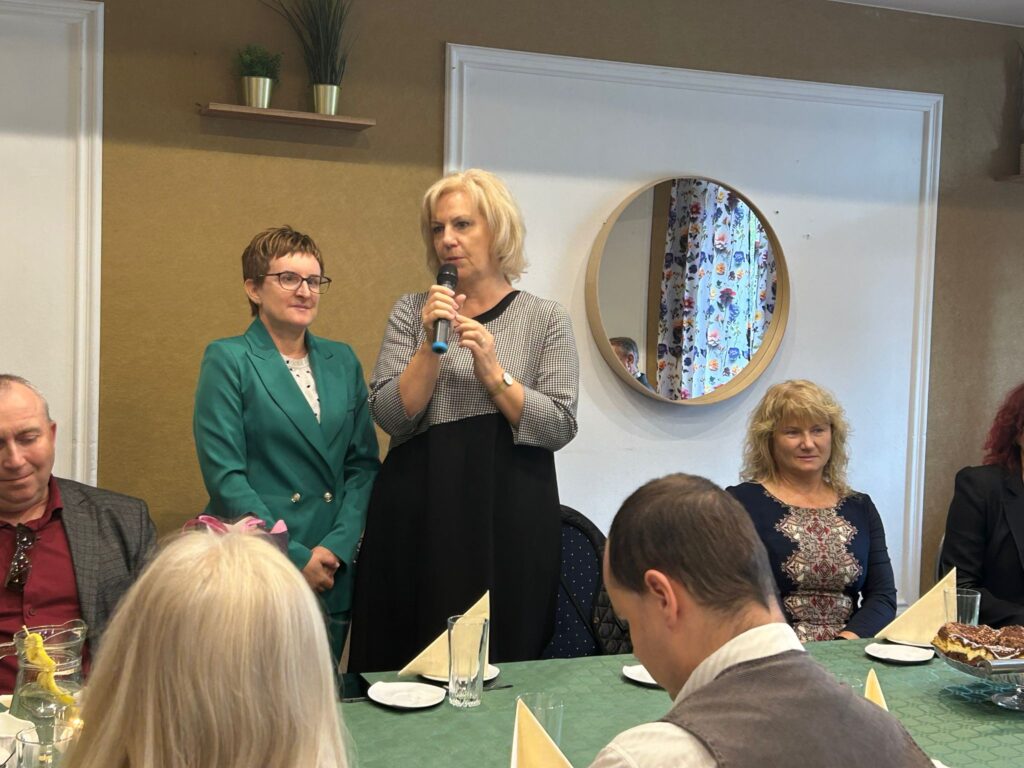 Na zdjęciu widać dwie kobiety stojące przy stole w restauracji „Kociewianka” podczas uroczystości z okazji Dnia Białej Laski. Jedna z kobiet trzyma mikrofon i przemawia do zgromadzonych, ubrana w czarną sukienkę, a obok niej stoi druga kobieta w zielonym garniturze, która słucha. W tle widoczna jest okrągła lustrzanka na ścianie, odbijająca fragment wnętrza restauracji, z kwiecistą zasłoną. W pierwszym planie siedzą goście przy stole, część z nich patrzy w stronę mówiącej. Na stole widoczna jest herbata, napój oraz kawałki ciasta. Cała scena ma przyjazną, uroczystą atmosferę.