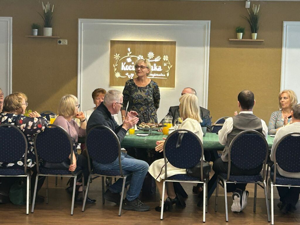 Na zdjęciu widać grupę osób siedzących przy stole w restauracji „Kociewianka” w Starogardzie Gdańskim podczas uroczystości z okazji Dnia Białej Laski. W centrum zdjęcia stoi kobieta w kolorowej sukience, przemawiająca lub dziękująca za występ. Osoby przy stole, w tym kobiety i mężczyźni, patrzą na nią i aplauzują, niektórzy klaszczą. Na stole stoją szklanki z napojami, talerze oraz ciasto. W tle widoczna jest ozdobna tablica z napisem „Kociewianka”, a cała scena ma ciepłą, serdeczną atmosferę.