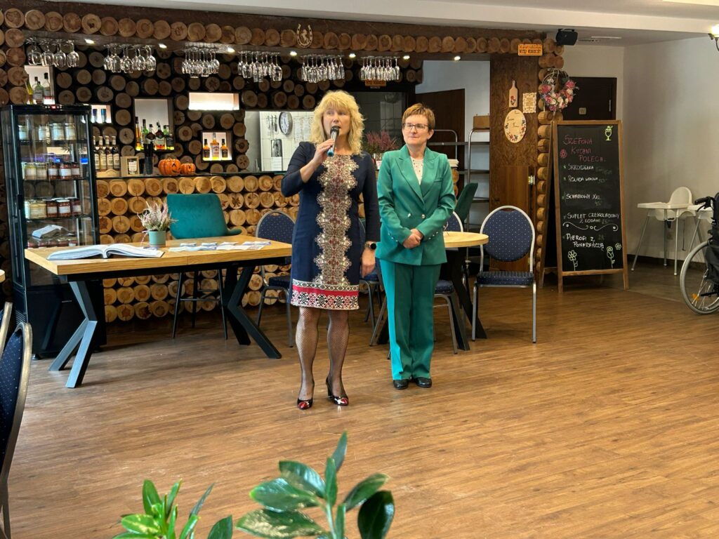 Zdjęcie przedstawia dwie kobiety stojące w restauracji „Kociewianka” podczas uroczystości z okazji Dnia Białej Laski. Pierwsza kobieta, ubrana w elegancką, ciemną sukienkę z haftami, trzyma mikrofon i przemawia do zgromadzonych. Obok niej stoi druga kobieta, w zielonym garniturze, która słucha i patrzy na przemawiającą. W tle widać stylowe wnętrze restauracji, z drewnianymi elementami dekoracyjnymi, takim jak ściana z okrągłych desek i wieszak na szklanki. W rogu znajduje się tablica informacyjna z ofertą restauracji, a cała scena ma ciepłą i przyjazną atmosferę.