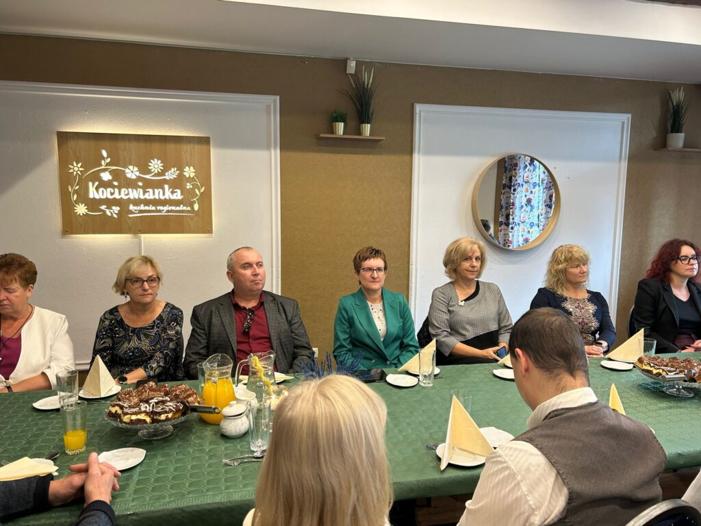 Zdjęcie przedstawia grupę osób siedzących przy stole w restauracji „Kociewianka” w Starogardzie Gdańskim, podczas uroczystości z okazji Dnia Białej Laski. W pierwszym planie widzimy kilka osób, w tym kobiety i mężczyzn, którzy patrzą przed siebie, uczestnicząc w spotkaniu. Na stole stoją napoje, talerze i ciasto. W tle widoczna jest ozdobna tablica z napisem „Kociewianka” oraz okrągłe lustro, które odbija fragment wnętrza. Cała scena ma przyjazną atmosferę, a osoby obecne na spotkaniu wydają się być zaangażowane w rozmowy i wydarzenia.