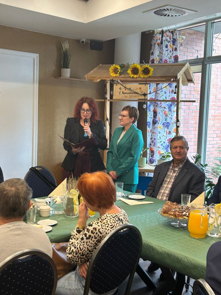 Na zdjęciu widzimy moment z uroczystości z okazji Dnia Białej Laski. Dwie kobiety stoją przy mikrofonie – jedna z nich trzyma dokumenty i przemawia, a druga, ubrana w zielony garnitur, słucha z uśmiechem. W tle widoczny jest dekoracyjny namiot z napisem „Kociewianka” ozdobiony słonecznikami. Przy stole siedzą goście, w tym mężczyzna w eleganckim garniturze i kobieta w bluzce w leopardzki wzór, która słucha przemówienia. Na stole widoczna jest lemoniada i ciasta, a w tle przez okno widać zieleń roślin. Cała scena emanuje serdeczną i uroczystą atmosferą.