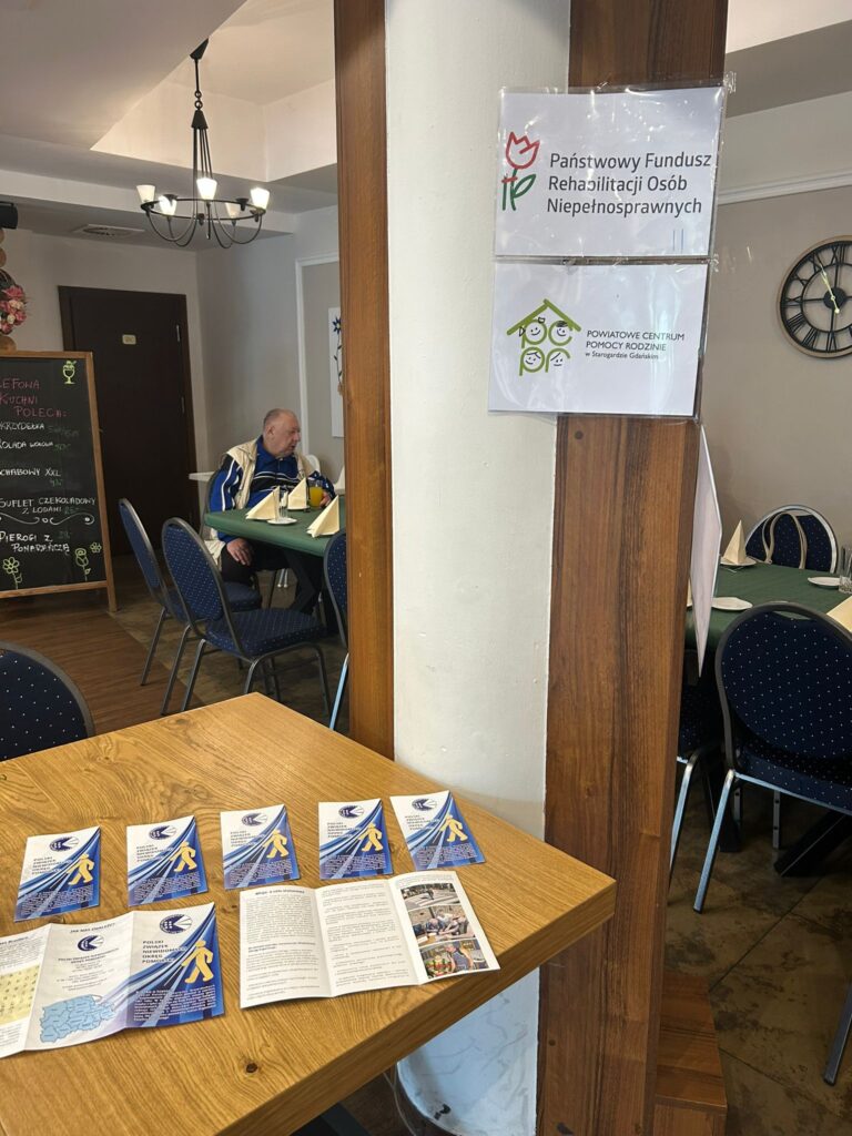 Zdjęcie przedstawia stół w restauracji „Kociewianka” w Starogardzie Gdańskim, na którym leżą ulotki informacyjne dotyczące Państwowego Funduszu Rehabilitacji Osób Niepełnosprawnych oraz Powiatowego Centrum Pomocy Rodzinie w Starogardzie Gdańskim. Na stole widocznych jest kilka broszur, a w tle, przy stole, siedzi starszy mężczyzna. W tle widać także tablicę informacyjną z logo obu instytucji, zawieszoną na filarze w lokalu. Cała scena odbywa się w przestronnym pomieszczeniu z eleganckimi detalami, takim jak drewniana podłoga i klasyczne oświetlenie.