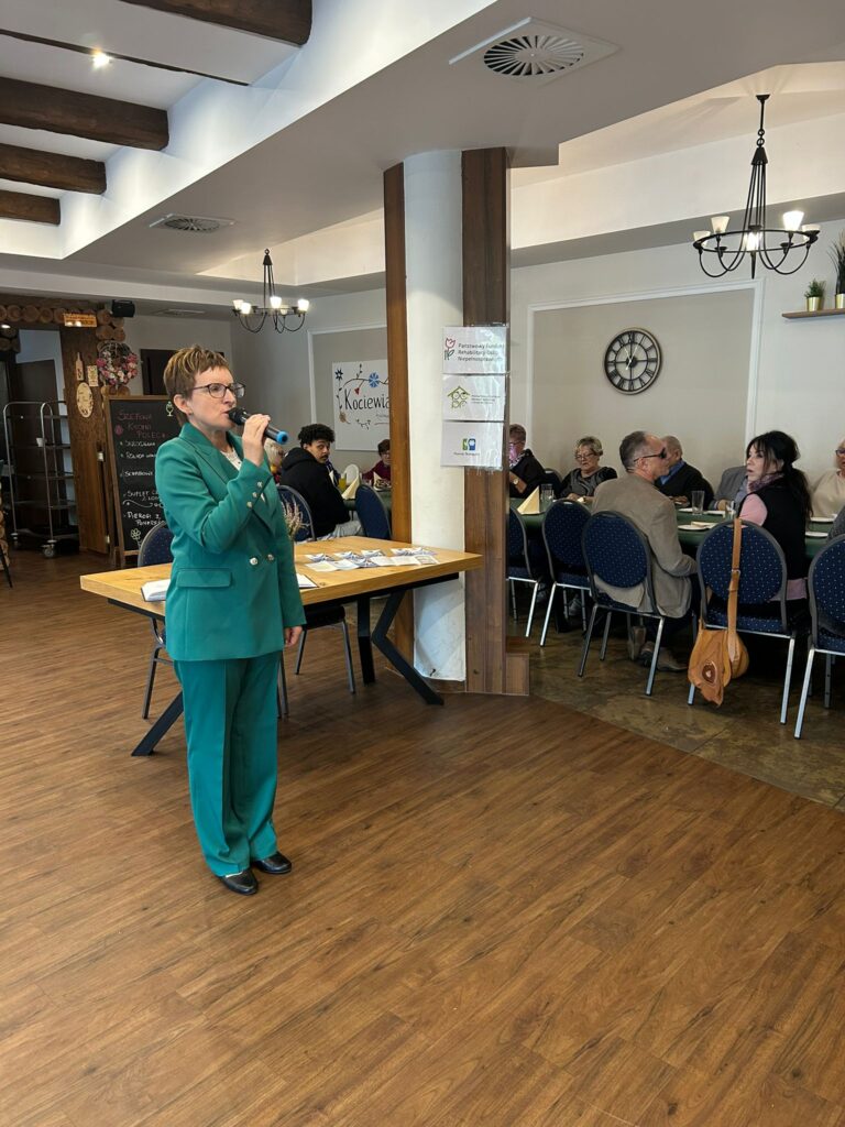 Zdjęcie przedstawia uroczystość z okazji Dnia Białej Laski 2025, która odbyła się w restauracji „Kociewianka” w Starogardzie Gdańskim. Na pierwszym planie stoi kobieta w zielonym garniturze, przemawiająca do mikrofonu. W tle przy stołach siedzą członkowie i goście Koła, słuchając wystąpień. Pomieszczenie jest przestronne, z jasnymi ścianami, drewnianą podłogą i ozdobnymi żyrandolami. Panuje uroczysta i życzliwa atmosfera, a spotkanie umożliwiło wspólne świętowanie, wymianę informacji oraz integrację osób niewidomych z całego regionu.