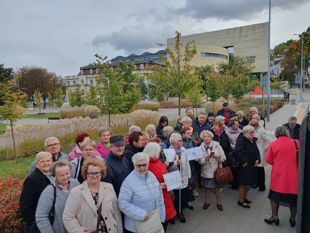 Grupa osób starszych stojących razem na chodniku przed Teatrem Muzycznym w Gdyni. Uczestnicy wycieczki ubrani są w jesienne ubrania, w tle widoczny jest nowoczesny budynek teatru, zieleń miejska i pochmurne niebo. Niektórzy trzymają tabliczki i kartki. Atmosfera wspólnego spotkania.