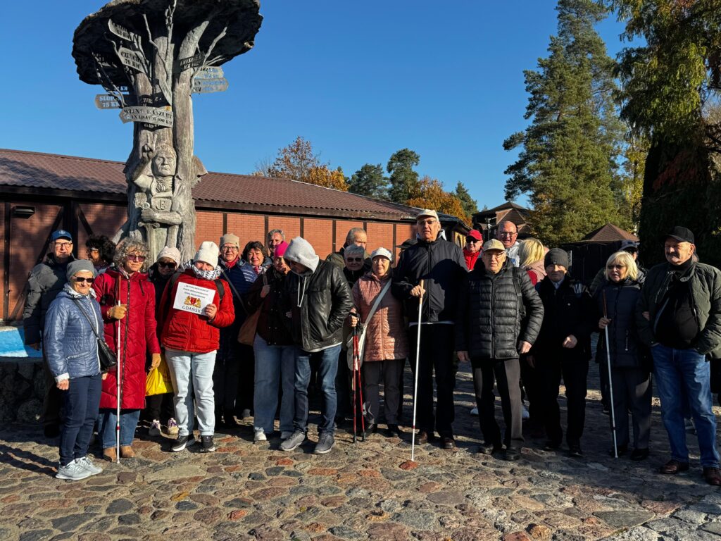Grupa uczestników projektu „Aktywni Niewidomi IV” podczas wydarzenia plenerowego w jesiennej scenerii. Osoby z dysfunkcją wzroku wraz z przewodnikami stoją na brukowanym placu przed pomnikiem w kształcie drzewa z rzeźbionymi postaciami i kierunkowskazami. Uczestnicy ubrani są w kurtki, czapki i okulary przeciwsłoneczne, co wskazuje na chłodniejszą pogodę. W tle widoczne są budynki z brązowymi dachami oraz kolorowe liście drzew.