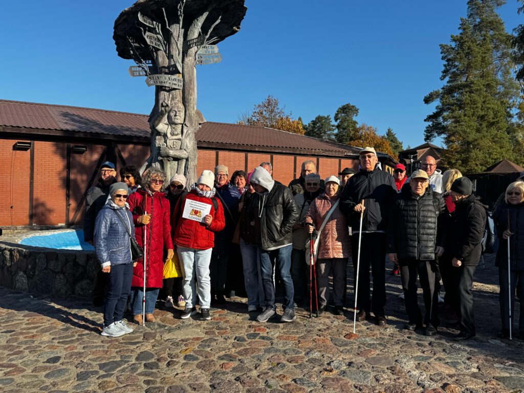 „Grupa uczestników projektu „Aktywni Niewidomi IV” zgromadzona przed kamienną rzeźbą przypominającą drzewo z kierunkowskazami i postaciami. Osoby z dysfunkcją wzroku, ubrane w ciepłą odzież, stoją na brukowanym placu obok niebieskiej fontanny. W tle widoczne są budynki z cegły i drzewa w jesiennej szacie. Jedna osoba trzyma tabliczkę z logo wydarzenia.”