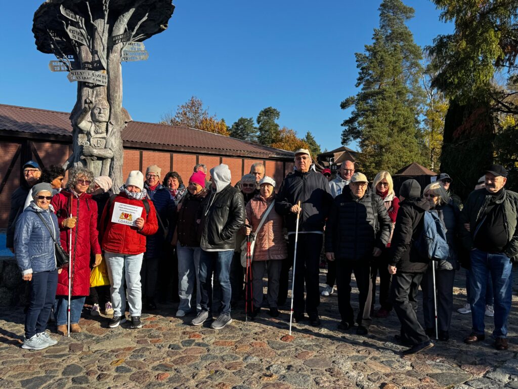 Uczestnicy projektu „Aktywni Niewidomi IV” podczas jesiennego wyjazdu plenerowego, stojący na brukowanej ścieżce przed drewnianą rzeźbą z kierunkowskazami. Osoby z dysfunkcją wzroku wraz z przewodnikami pozują wspólnie w chłodnej, słonecznej aurze. W tle widoczne są ceglane budynki i kolorowe liście drzew.