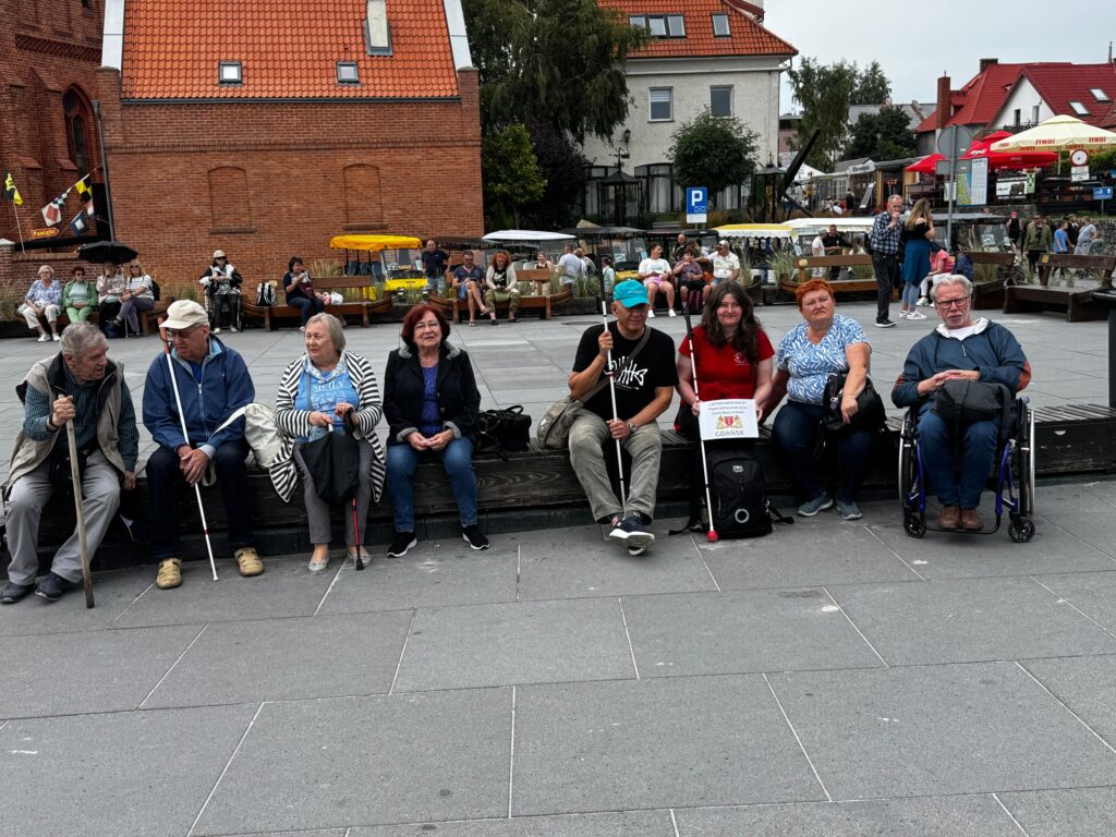 Uczestnicy projektu „Aktywni Niewidomi IV” siedzą na drewnianym podwyższeniu na rynku miejskim, otoczeni przez przechodniów i kawiarniane stoliki. Wśród nich są osoby z niepełnosprawnością wzrokową, korzystające z lasek i wózka inwalidzkiego. Jedna osoba trzyma mikrofon, prowadząc wystąpienie lub występ. W tle widoczne są ceglane budynki, parasole i jesienne drzewa, tworzące tło dla integracyjnego wydarzenia w przestrzeni publicznej.