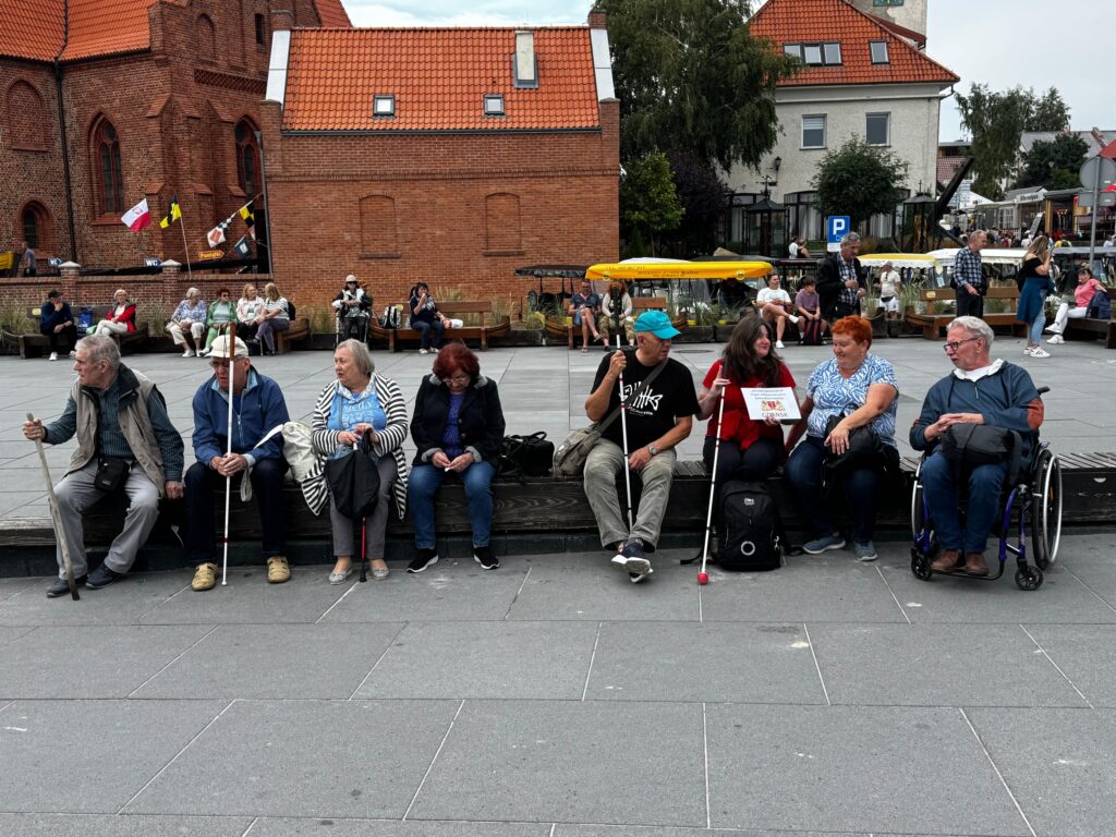 Grupa uczestników projektu „Aktywni Niewidomi IV” siedzi na drewnianym podwyższeniu na miejskim placu. Wśród nich są osoby z dysfunkcją wzroku, trzymające białe laski, oraz osoba na wózku inwalidzkim. W tle widoczne są ceglane budynki, flagi i przechodnie, co tworzy atmosferę otwartego, dostępnego miejsca publicznego.