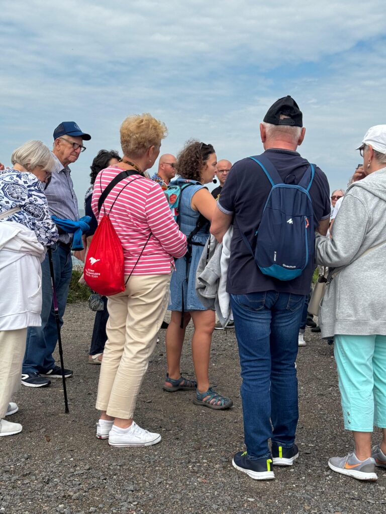 Uczestnicy projektu „Aktywni Niewidomi IV” zgromadzeni na żwirowej ścieżce podczas wyjazdu integracyjnego. Osoby z dysfunkcją wzroku oraz ich przewodnicy słuchają kobiety w niebieskiej sukience, która prowadzi spotkanie. W tle widoczne jest niebo z lekkim zachmurzeniem, a uczestnicy ubrani są w codzienne stroje, niektórzy z plecakami i nakryciami głowy.