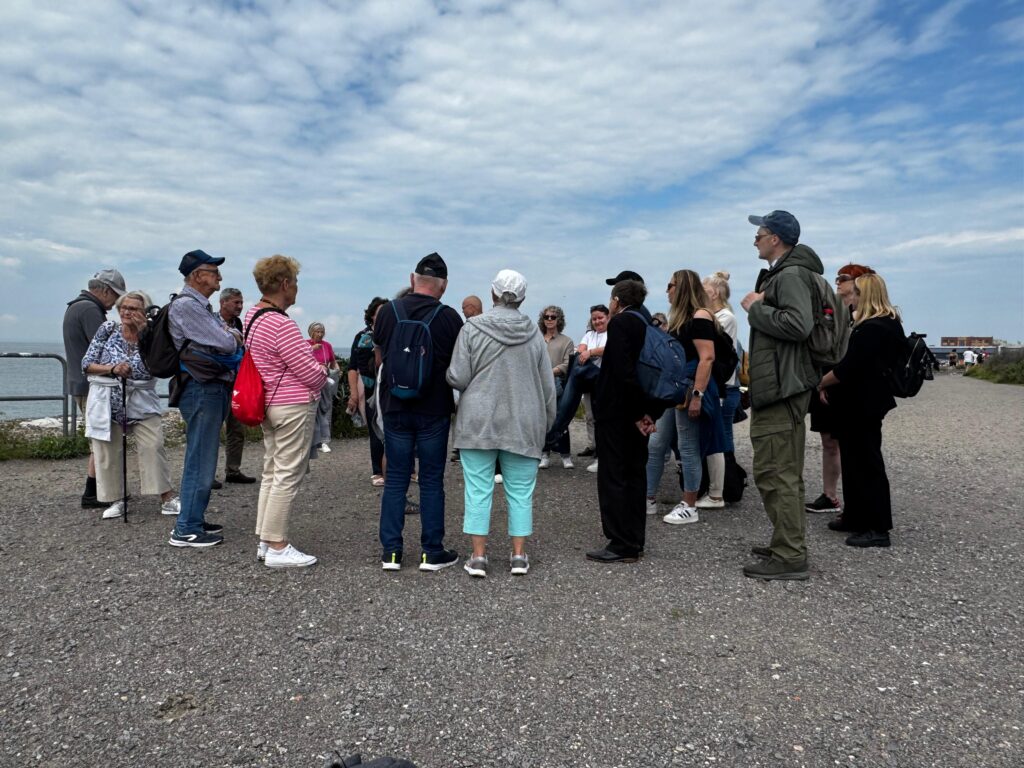 Uczestnicy projektu „Aktywni Niewidomi IV” podczas wyjazdu integracyjnego spacerują żwirową ścieżką w pobliżu zbiornika wodnego. Grupa słucha przewodnika, a osoby z dysfunkcją wzroku oraz ich opiekunowie ubrani są w codzienne stroje, z plecakami i nakryciami głowy. W tle widoczne jest częściowo zachmurzone niebo i spokojna, nadmorska sceneria.