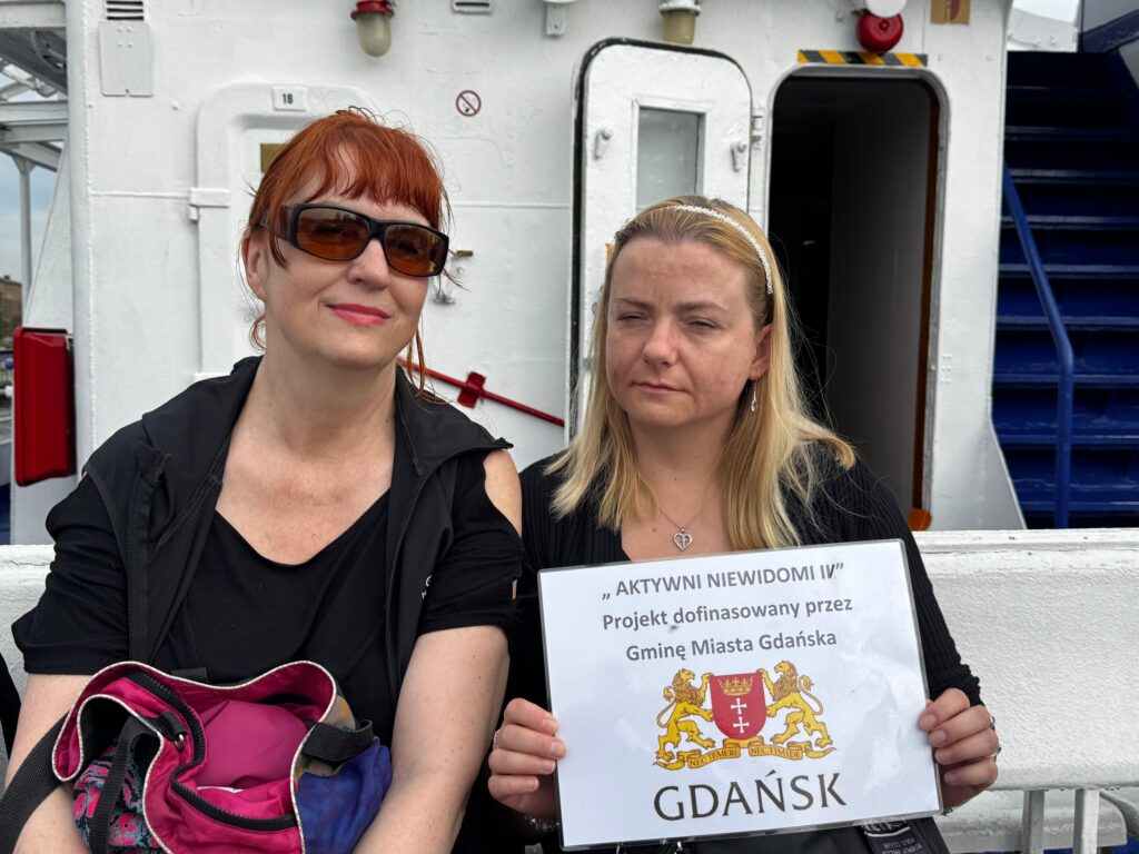 Dwóch uczestników projektu „Aktywni Niewidomi IV” siedzi na ławce na pokładzie promu. Jeden z nich trzyma tabliczkę z nazwą projektu i herbem Gdańska. W tle widoczne są elementy statku, takie jak drzwi, schody i sprzęt ratunkowy, co wskazuje na morską scenerię wycieczki.