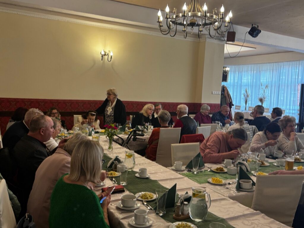 Uczestnicy obchodów Międzynarodowego Dnia Białej Laski siedzą przy elegancko nakrytych stołach w restauracji Hacjenda w Gdańsku Wrzeszczu. Stoły udekorowane są białymi obrusami, zielonymi serwetkami i kwiatowymi kompozycjami. W tle widoczne są żyrandole i kinkiety, a goście – osoby z dysfunkcją wzroku oraz ich przewodnicy – rozmawiają i delektują się poczęstunkiem w serdecznej atmosferze.