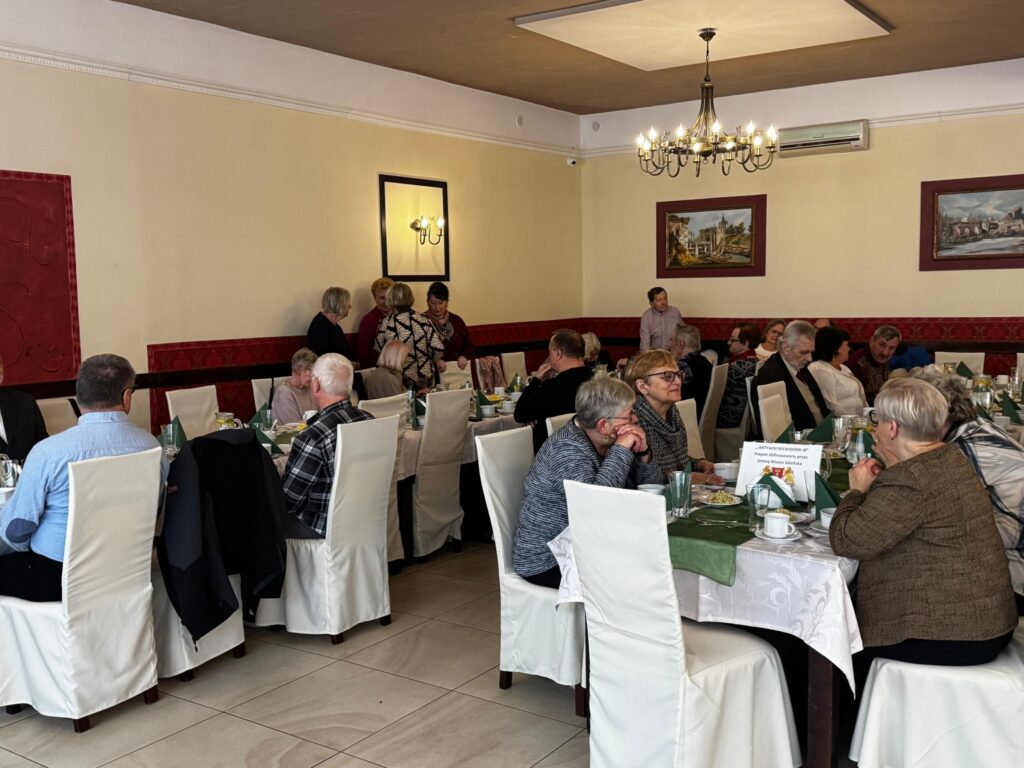 Seniorzy – uczestnicy obchodów Międzynarodowego Dnia Białej Laski – siedzą przy elegancko nakrytych stołach w restauracji Hacjenda w Gdańsku Wrzeszczu. Stoły ozdobione są białymi obrusami, zielonymi bieżnikami i szklanymi naczyniami. Na ścianach wiszą obrazy, a z sufitu zwisają żyrandole. Goście rozmawiają w przyjaznej atmosferze, ciesząc się wspólnym spotkaniem.