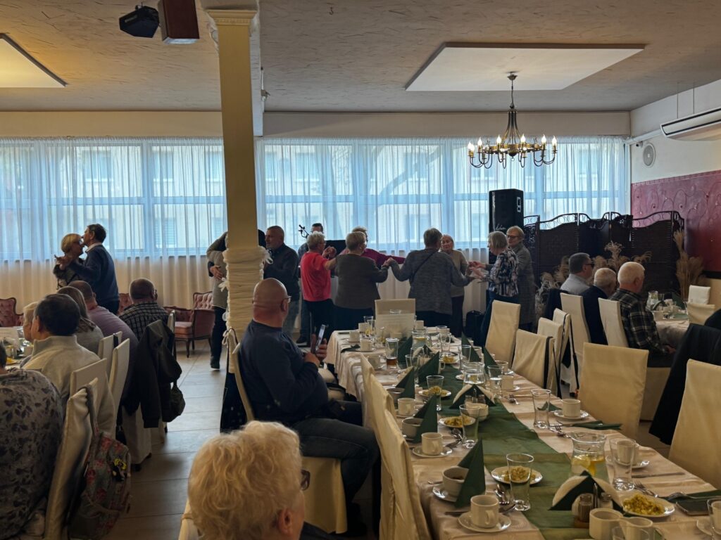 Uczestnicy obchodów Międzynarodowego Dnia Białej Laski tańczą i bawią się w jasnej sali restauracji Hacjenda w Gdańsku Wrzeszczu. Przy długim stole siedzą goście, a w tle widoczne są duże okna z białymi firanami, żyrandol oraz sprzęt muzyczny. Radosna atmosfera podkreśla integracyjnharakter spotkania osób z dysfunkcją wzroku i ich przewodników.