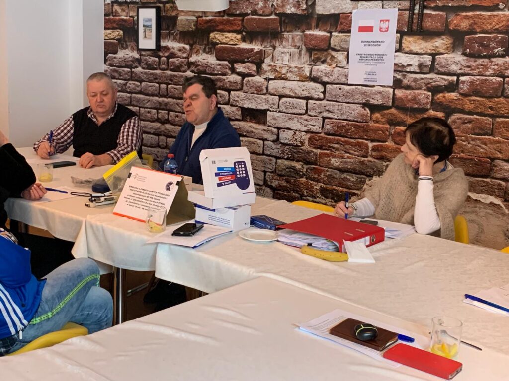 Grupa czterech osób siedzi przy stole podczas szkolenia. Na stole leżą dokumenty, długopisy, telefon, szklanka z napojem, banan, czerwony segregator i kilka pudełek. W tle znajduje się ściana z ceglanej tapety oraz plakat z informacją o dofinansowaniu ze środków PFRON.