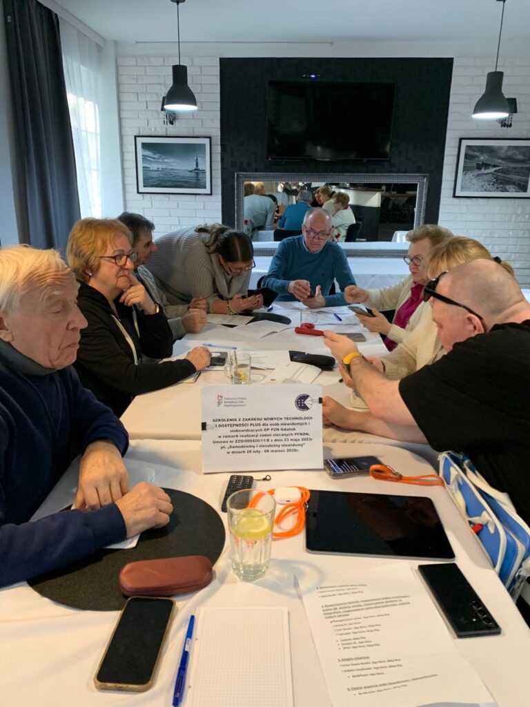 Grupa osób siedzi przy prostokątnym stole podczas szkolenia. Uczestnicy korzystają ze smartfonów, tabletów i materiałów drukowanych. Na stole stoją szklanki z wodą, długopisy, notatki i kartka z informacją o szkoleniu dotyczącym nowych technologii i dostępności plus. W tle widać duże lustro, telewizor na ścianie oraz czarno-białe zdjęcia przedstawiające nadmorskie krajobrazy.
