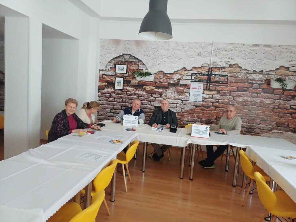 Pięć osób siedzi przy stołach nakrytych białymi obrusami w jasnym pomieszczeniu z drewnianą podłogą i żółtymi krzesłami. Na stołach znajdują się dokumenty, talerzyki z przekąskami oraz tabliczki informacyjne. W tle widać ścianę z tapetą imitującą cegłę, ozdobioną ramkami i roślinami. Uczestnicy rozmawiają i biorą udział w spotkaniu lub szkoleniu.