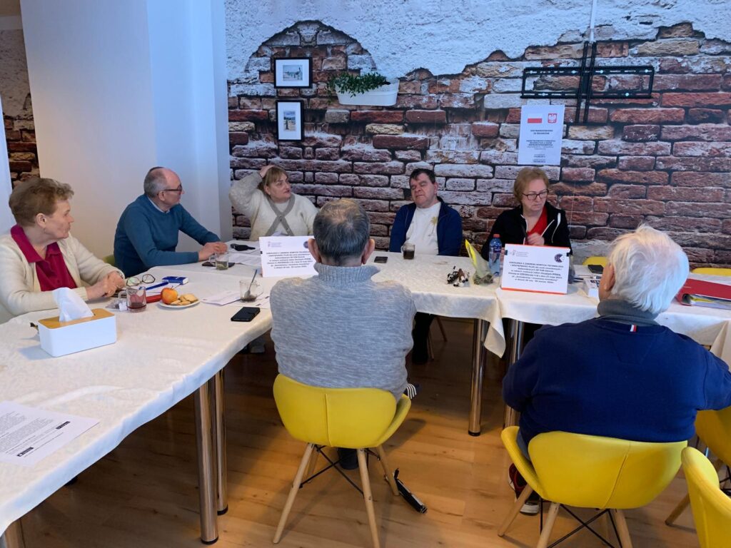 Grupa siedmiu starszych osób siedzi przy dwóch stołach nakrytych białymi obrusami w jasnym pomieszczeniu. Na stołach leżą dokumenty, notesy, długopisy, okulary, owoce, napoje oraz pudełko chusteczek. W tle znajduje się ściana z tapetą imitującą cegłę i dwoma małymi obrazkami. Na jednym ze stołów stoją dwie tabliczki informacyjne dotyczące projektu dla seniorów.