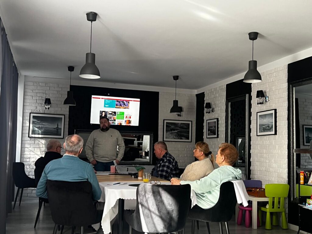 Grupa sześciu osób siedzi przy stole w nowoczesnym, jasnym pomieszczeniu. Jedna osoba stoi obok dużego ekranu na ścianie i prezentuje treści wyświetlone na stronie internetowej. Na ścianach wiszą czarno-białe zdjęcia, a w rogu znajduje się mały stolik dziecięcy z kolorowymi krzesełkami. Uczestnicy słuchają i patrzą w kierunku ekranu.