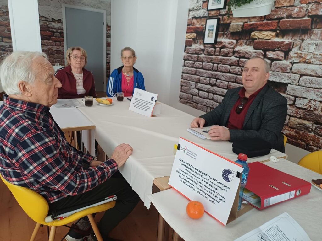 Cztery starsze osoby siedzą przy prostokątnym stole nakrytym białym obrusem w jasnym pomieszczeniu. Na stole leżą dokumenty, czerwony segregator, butelka wody, banan, pomarańcza oraz filiżanki z kawą. Mężczyzna po prawej stronie wygląda na prowadzącego spotkanie, a pozostali uczestnicy słuchają. W tle znajduje się ściana z tapetą imitującą cegłę oraz dwie tabliczki informacyjne dotyczące szkolenia.