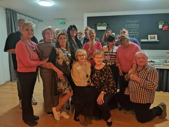 Około szesnaście osób pozuje wspólnie do zdjęcia w jasnym pomieszczeniu z drewnianą podłogą. Część osób stoi, a część klęczy z przodu. W tle znajduje się ciemna ściana z półkami, na których stoją rośliny i ramki, oraz blat z talerzami i jedzeniem. Na ścianie widoczny jest napis „Smacznego! Bon Appetit Guten Appetit Buon Appetito Provecho”.