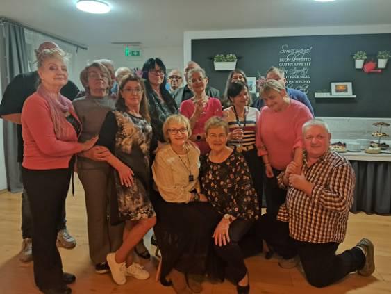 Grupa około piętnastu osób pozuje wspólnie do zdjęcia w sali z drewnianą podłogą. Część osób stoi, a część siedzi z przodu. W tle znajduje się stół z talerzami i przekąskami oraz ciemna ściana z małymi roślinami i ramkami. Oświetlenie jest ciepłe, tworząc przytulną atmosferę.