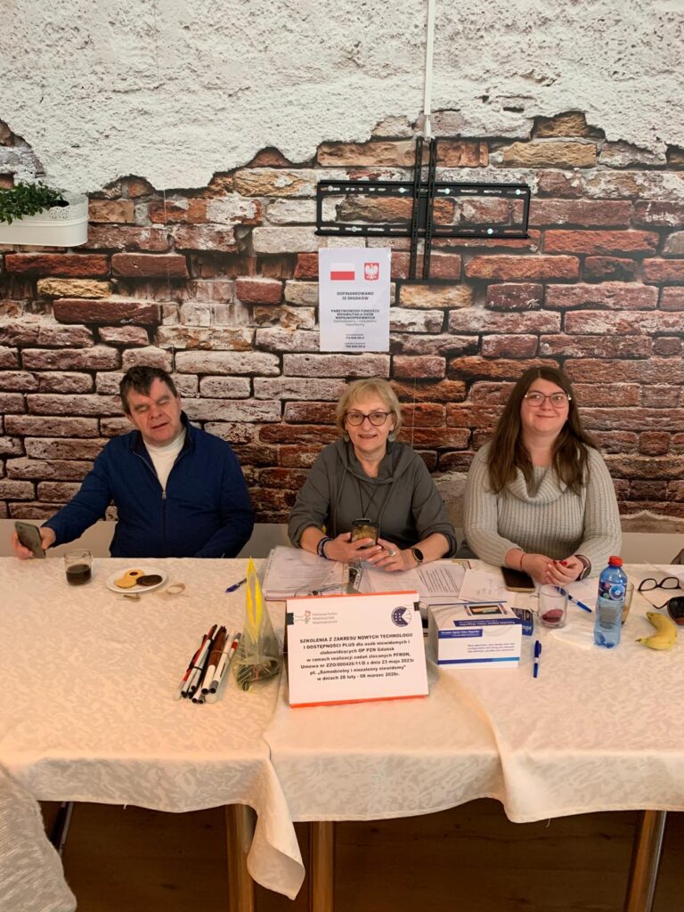 Trzy osoby siedzą przy stole nakrytym białym obrusem podczas szkolenia. Na stole leżą długopisy, teczki, dokumenty, pudełko, butelka wody, banan i drobne przekąski. W tle znajduje się ściana z tapetą imitującą cegłę oraz dwa plakaty, w tym jeden z informacją o dofinansowaniu ze środków PFRON.