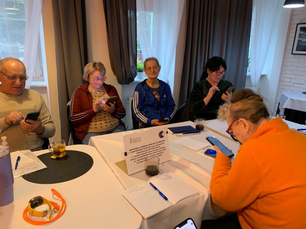 Kilka starszych osób siedzi przy stole w jasnym pomieszczeniu z dużymi oknami, korzystając ze smartfonów podczas zajęć. Na stole leżą notesy, długopisy, kubki z napojami oraz kartka z informacją o szkoleniu z nowych technologii dla osób 60+. Atmosfera jest spokojna i skupiona.
