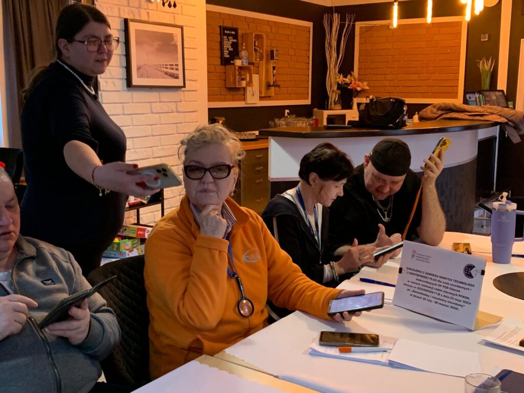 Pięć osób siedzi przy stole w ciepło oświetlonym pomieszczeniu, korzystając ze smartfonów i tabletów podczas szkolenia. Jedna osoba stoi i pokazuje coś na telefonie pozostałym uczestnikom. Na stole leżą notatki, dokumenty i kartka z informacją o szkoleniu z nowych technologii i dostępności dla osób 60 plus. W tle widać bar, półki i dekoracje.