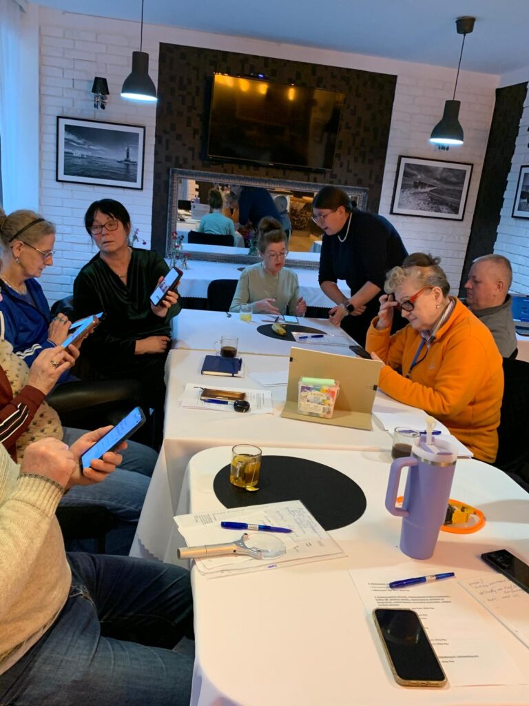 Pięć osób siedzi przy stole w ciepło oświetlonym pomieszczeniu, korzystając ze smartfonów i tabletów podczas szkolenia. Jedna osoba stoi i pokazuje coś na telefonie pozostałym uczestnikom. Na stole leżą notatki, dokumenty i kartka z informacją o szkoleniu z nowych technologii i dostępności dla osób 60 plus. W tle widać bar, półki i dekoracje.