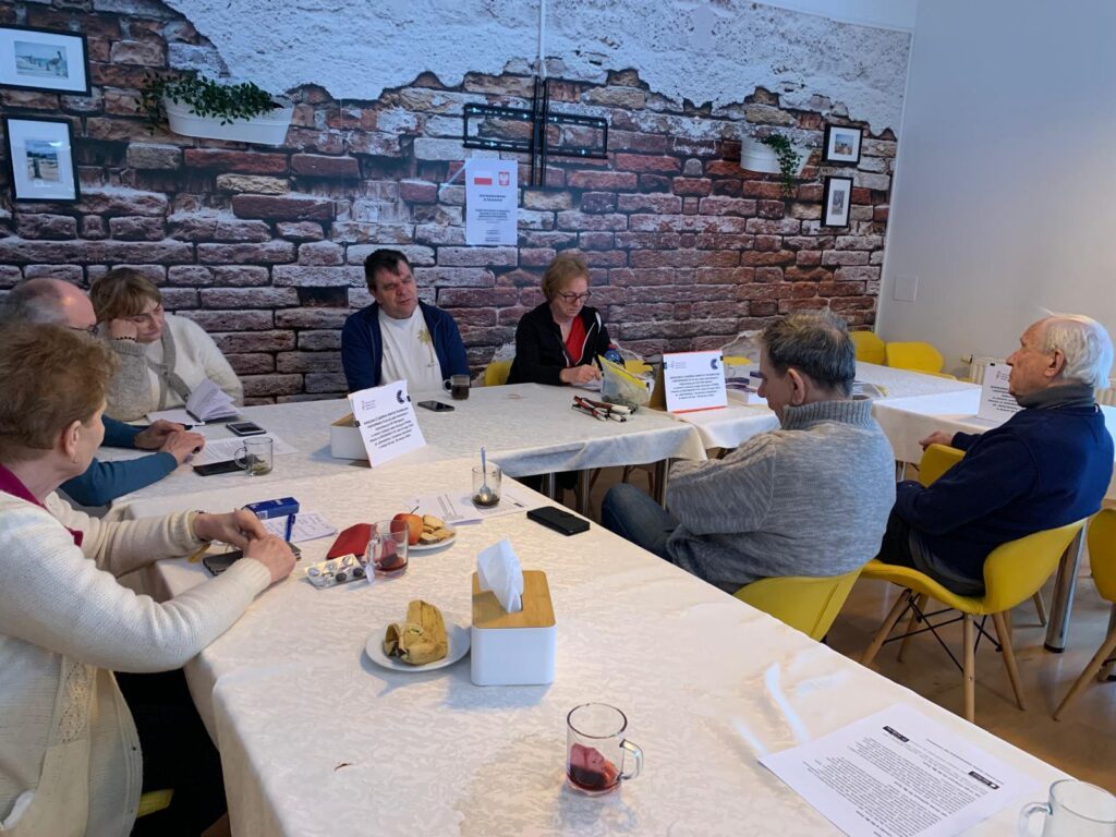 Grupa siedmiu osób siedzi przy dwóch stołach nakrytych białymi obrusami podczas spotkania lub szkolenia. Na stołach stoją talerzyki z przekąskami, napoje, dokumenty i tabliczki informacyjne. W tle znajduje się ściana z tapetą imitującą cegłę, ozdobiona ramkami i małymi roślinami. Uczestnicy rozmawiają i są zaangażowani w dyskusję.