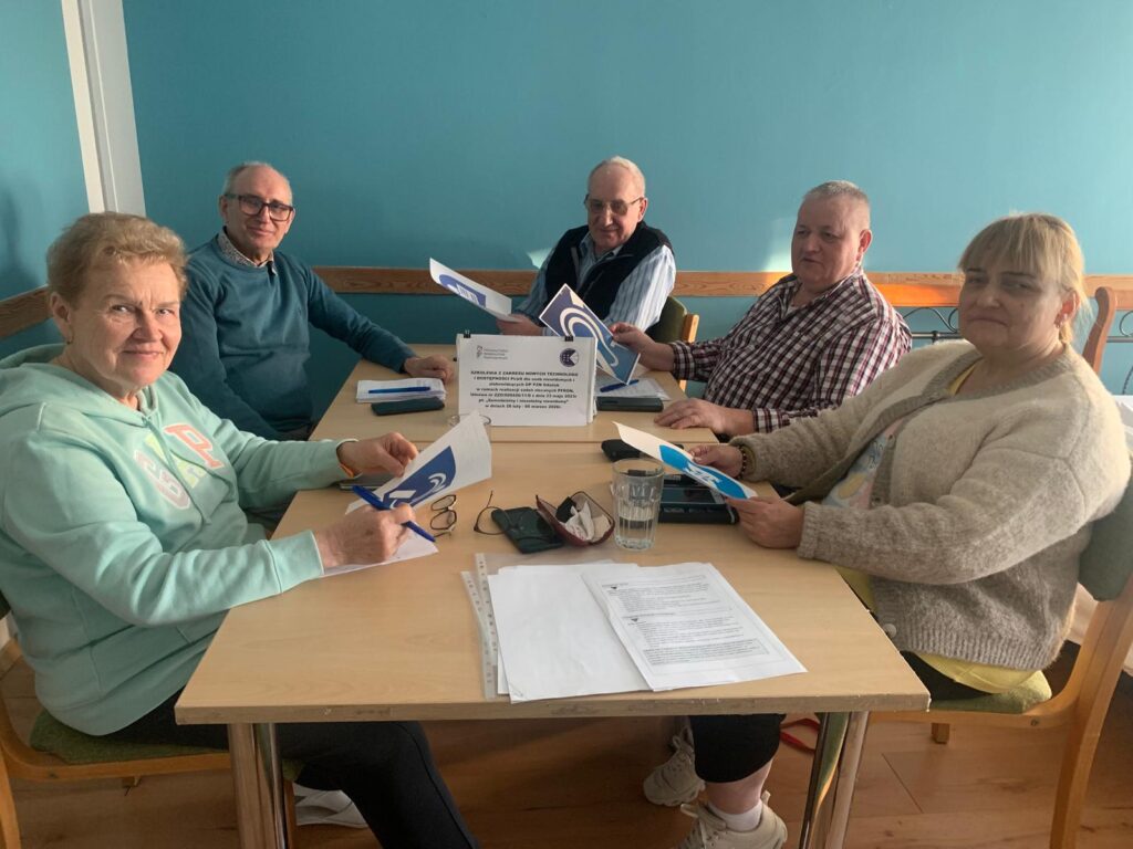 Pięć starszych osób siedzi przy drewnianym stole w jasnym pomieszczeniu o turkusowych ścianach. Każda osoba trzyma w rękach materiały drukowane. Na stole leżą dokumenty, szklanka z wodą i smartfon. Przed uczestnikami stoi kartka z informacją o szkoleniu dotyczącym obsługi komputera i internetu w ramach projektu „Aktywny Senior”.