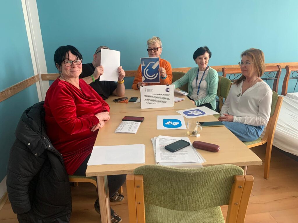 Pięć osób siedzi przy drewnianym stole w jasnym pomieszczeniu o błękitnych ścianach. Na stole leżą dokumenty, smartfon, okulary w etui oraz szklanka. Jedna osoba trzyma kartkę zasłaniającą twarz, inna trzyma niebieską tabliczkę z białym symbolem. Na środku stołu stoi kartka z informacją o szkoleniu dotyczącym wykorzystania technologii cyfrowych.